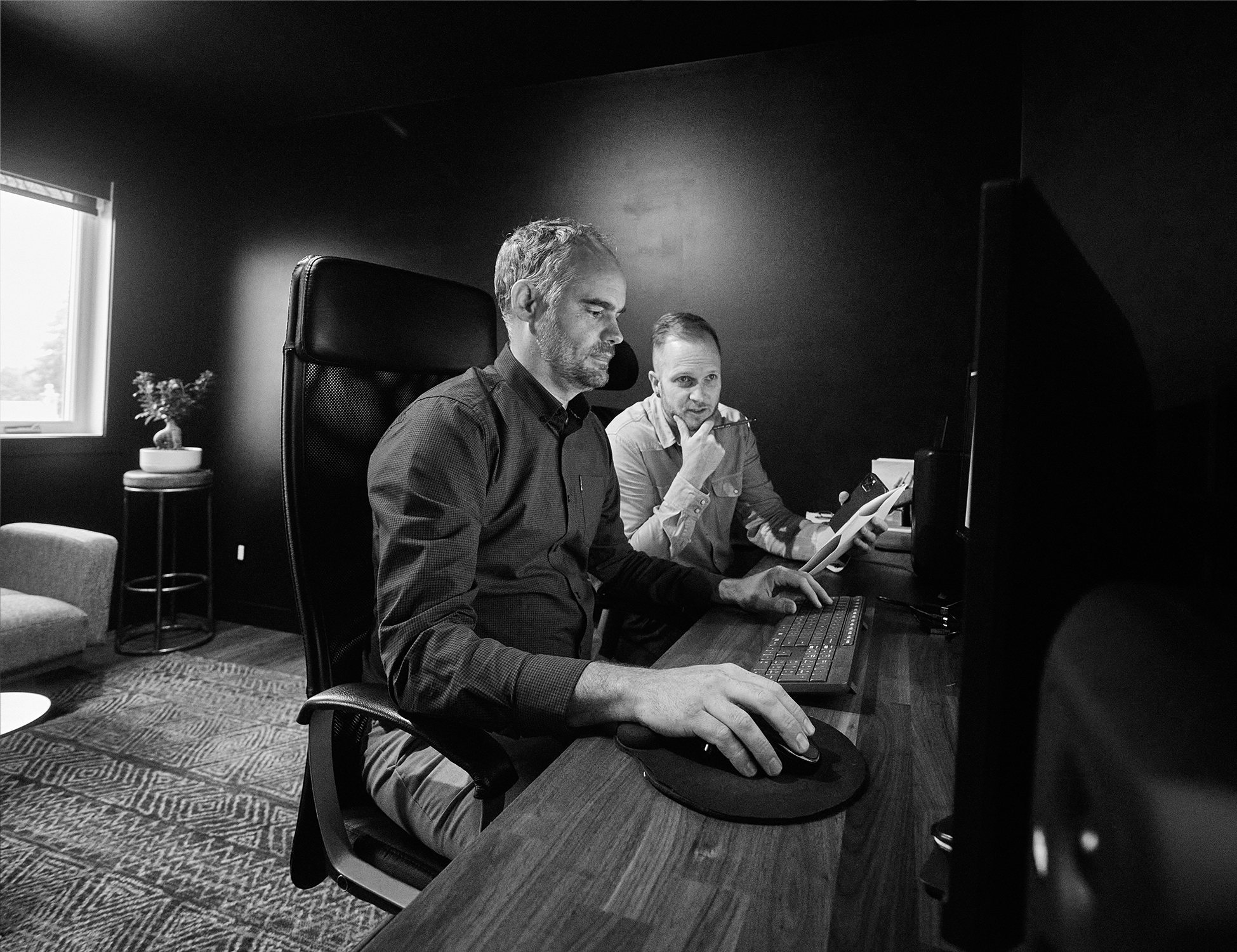 A black and white image of two Coelement team members working together at a desk with multiple monitors.