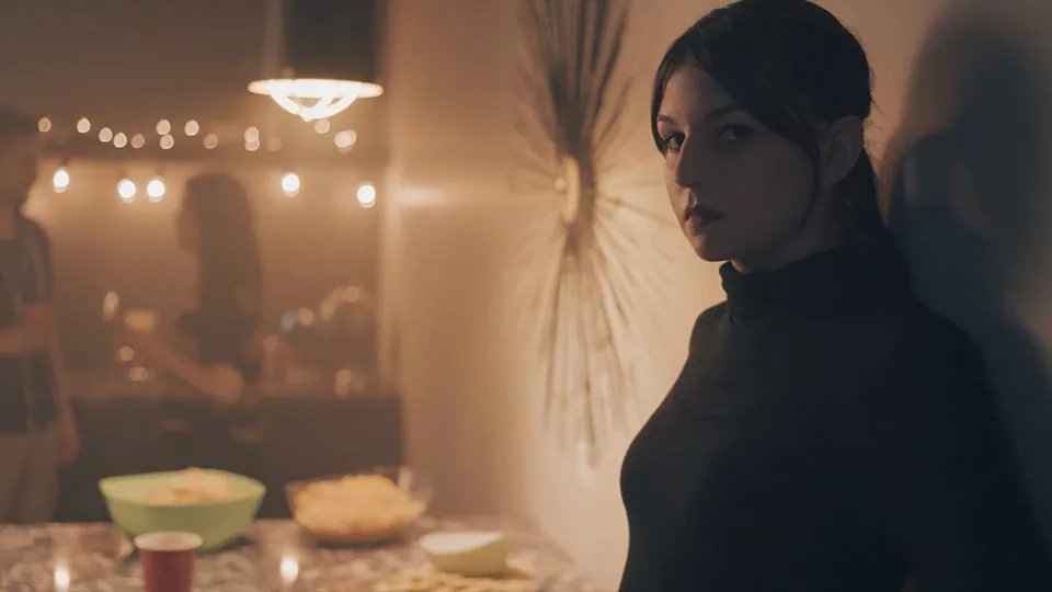 An image of a serious-faced women standing against a wall in a house party setting.
