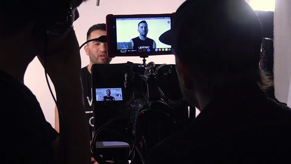 An image of a man behind a camera, filming a man talking against a white background, with the cameramen as a silhouettes.