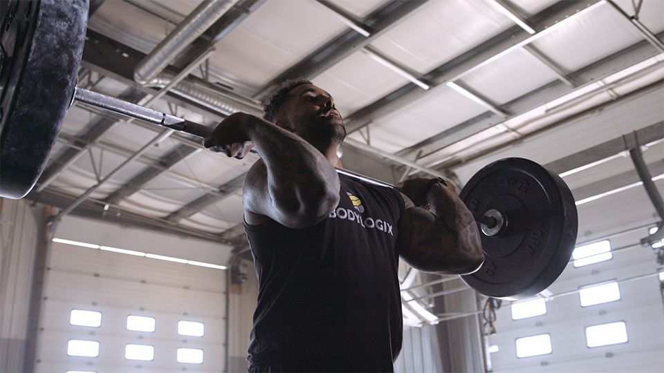 An image of a man lifting a barbell above his shoulders in a garage-like gym.
