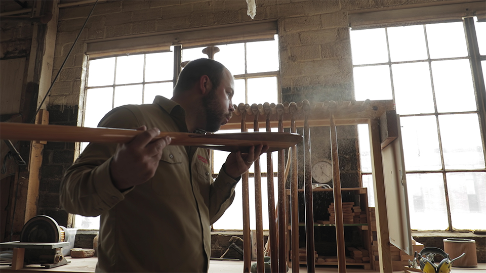 An image of a man blowing sawdust off of a canoe paddle his is crafting in a workshop.