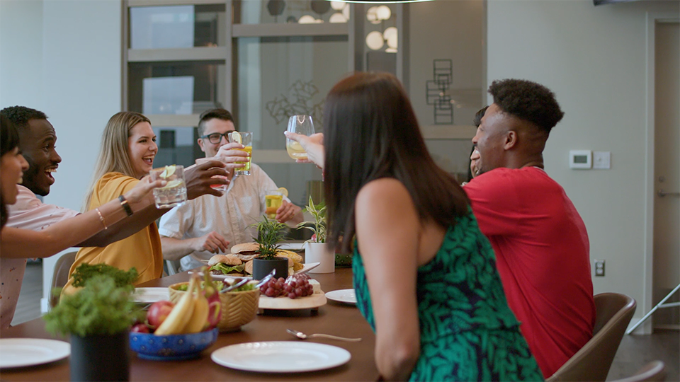 An image of a group of adults gathered around a dining table in a brightly lit room, smiling and participating in "cheers."