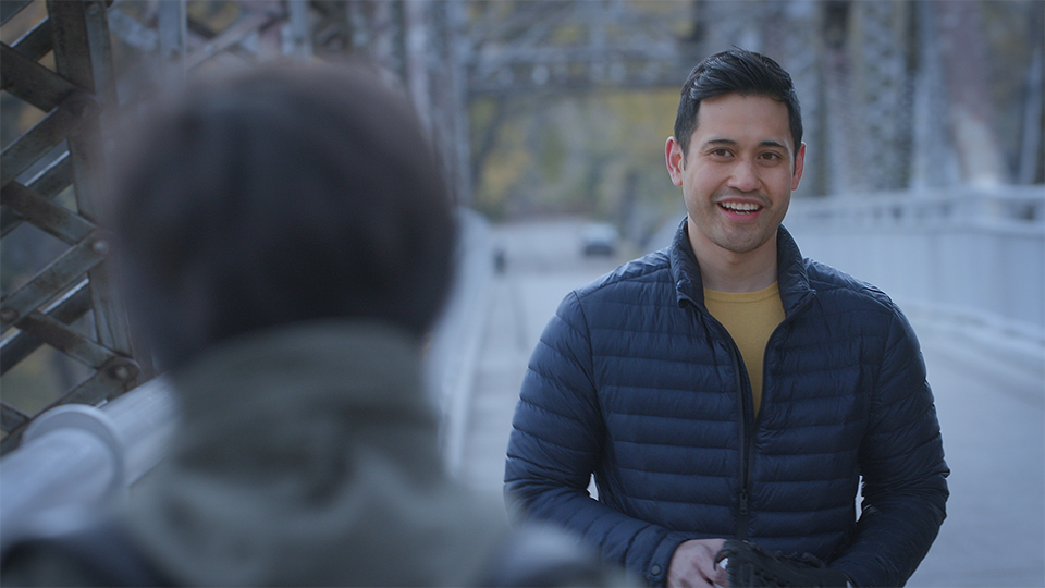 An image of a young man smiling and looking at an out of focus person standing in front of him. The people are standing on a bridge.