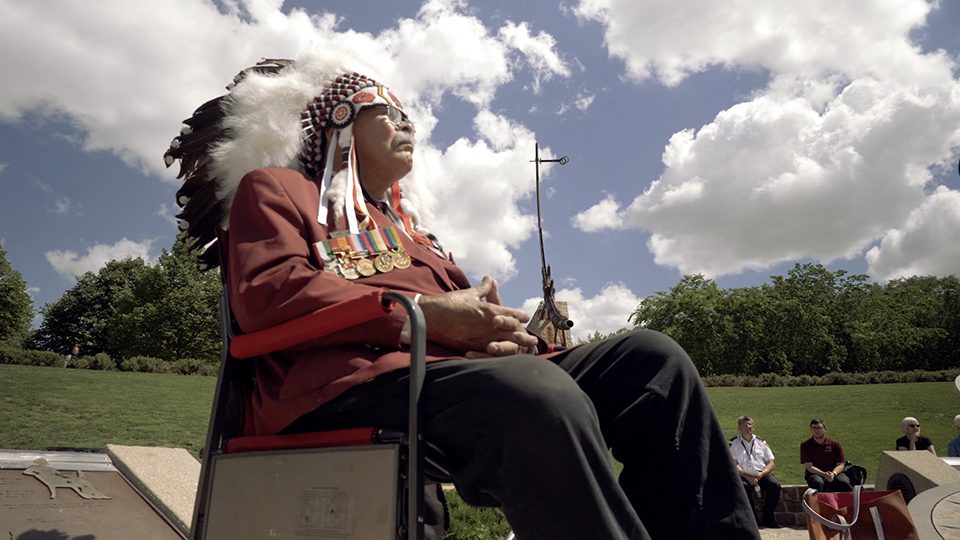 An image of an indigenous chief sitting on a chair in front of a group of people outside.