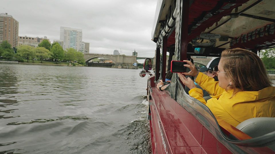 An image of a woman taking a photo of a river and the city of Boston from a tour boat.