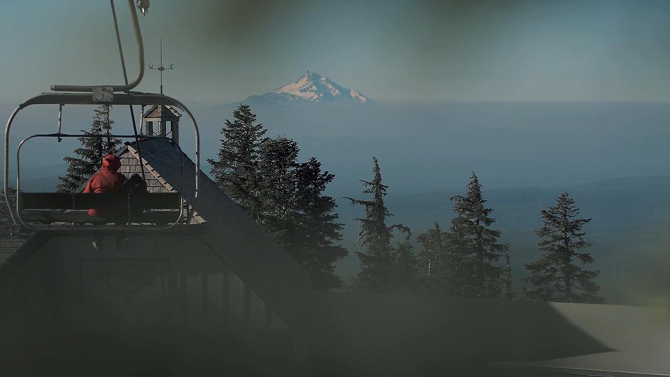 An image of person sitting on a ski lift with a lodge, tree-line and mountains in the background.