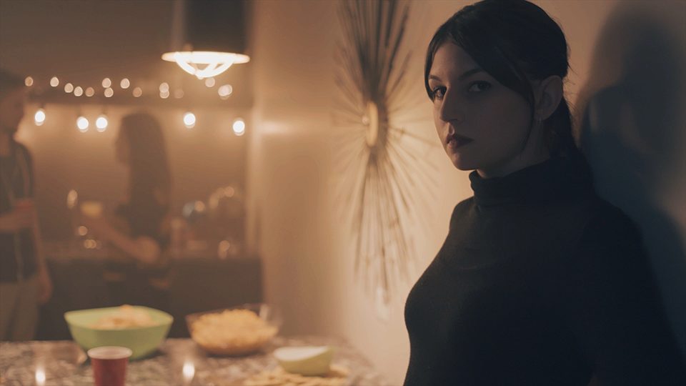 An image of a serious-faced women standing against a wall in a house party setting.