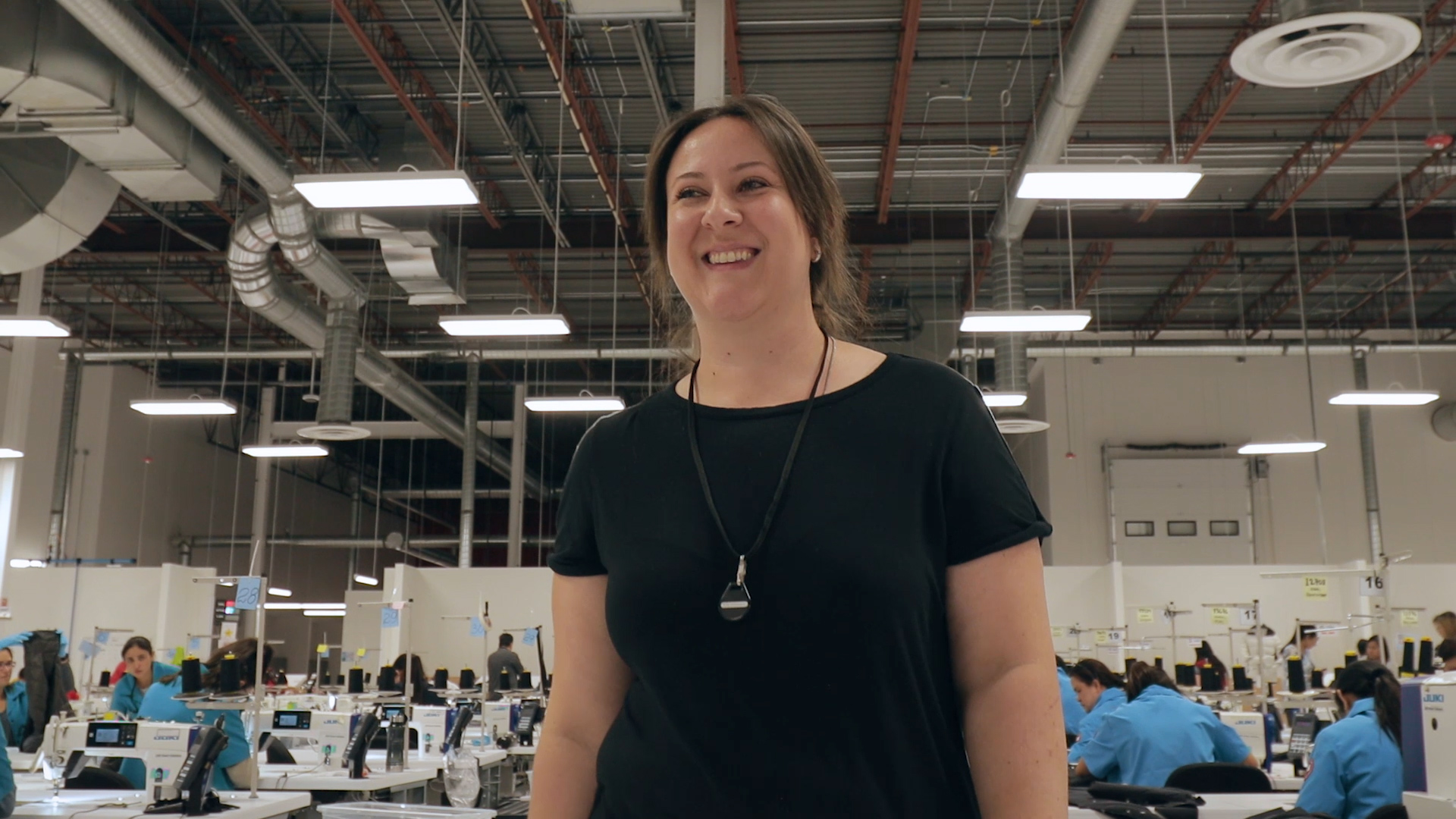 An image of a women smiling with a large group of people behind her working on sewing machines.