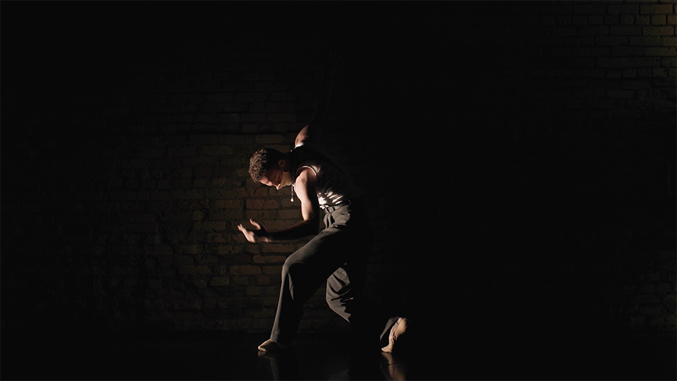 An image of a man in a bowed dance pose with his left arm bent and his head down. He is standing against a brick wall in a darkly lit room with a spotlight on his left side.