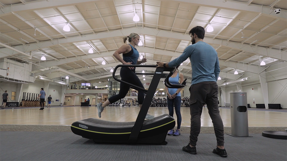 An image of a woman running on a treadmill with two trainers next to her. They are in a large gym with a large court and work out equipment behind them.