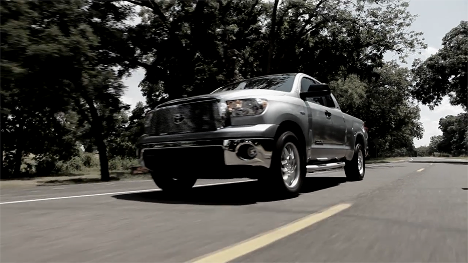 An image of a silver truck driving on a road, with a forest of trees in the background lining the road.