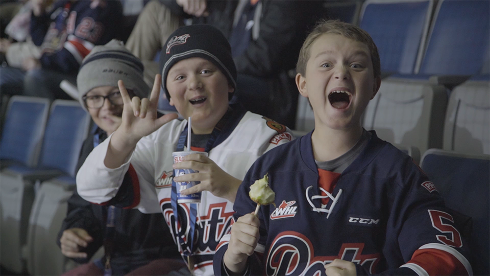 An image of three boys, cheering and smiling, facing the camera. They are sitting in stadium chairs at a hockey game and are wearing jerseys.