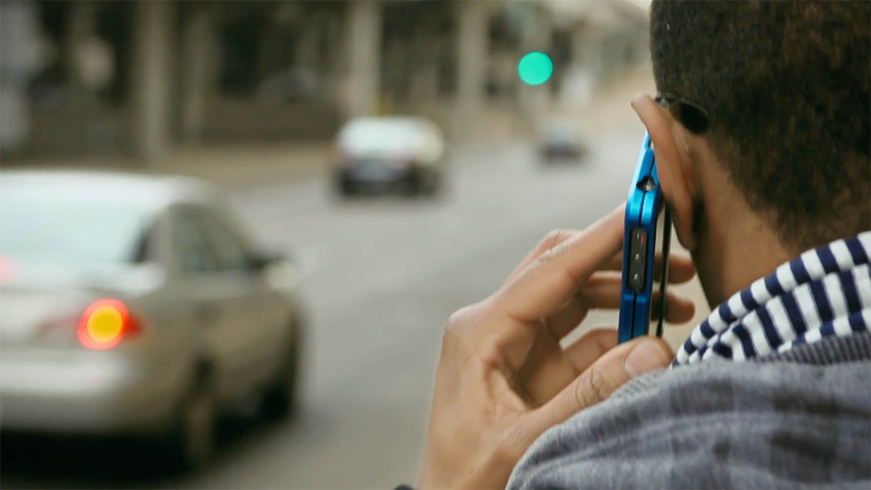 A close up image of a man holding a cell phone to his ear with a city street in front of him.