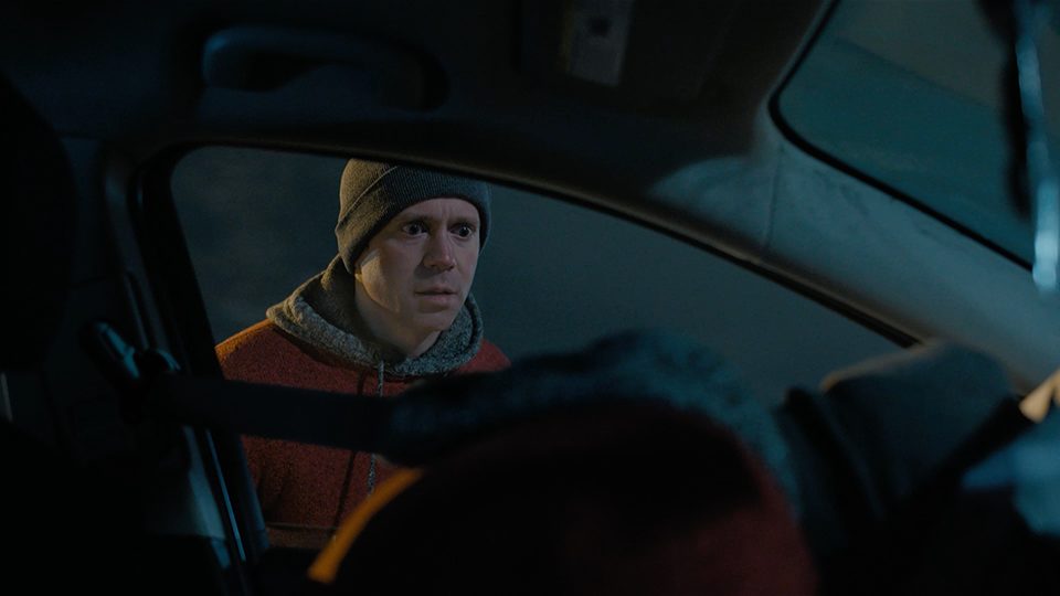 A frame from a live action Manitoba Public Insurance commercial, showing a man peering into a car window where he is lying against the steering wheel. The setting is at night and the man has a shocked expression on his face.